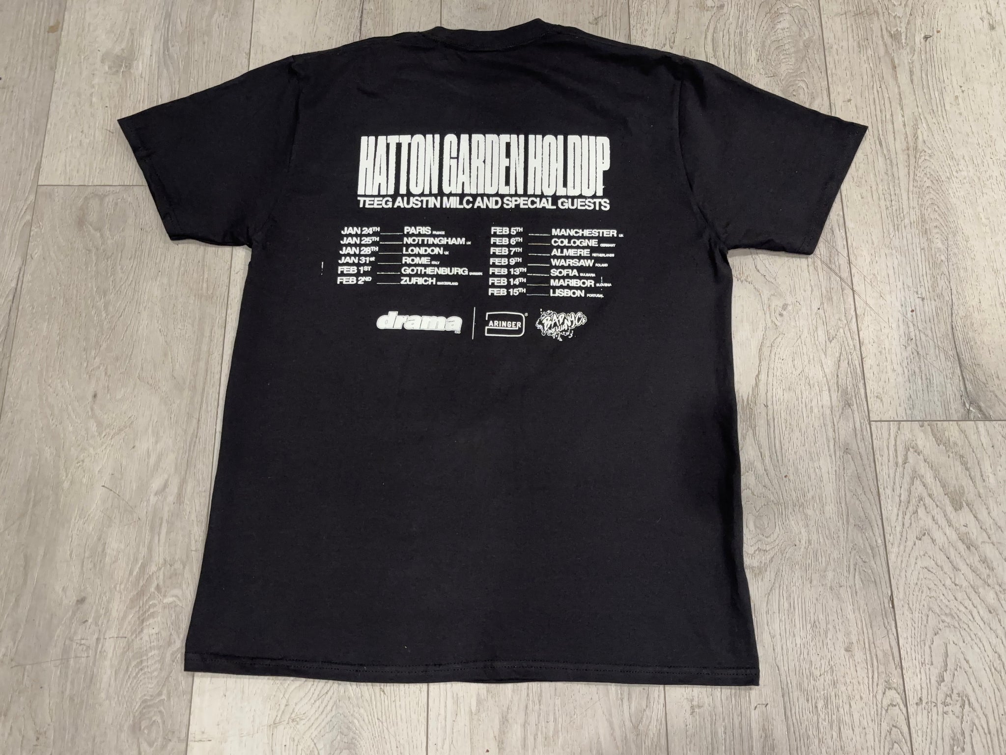 Black t-shirt with white text and logos on a wooden floor